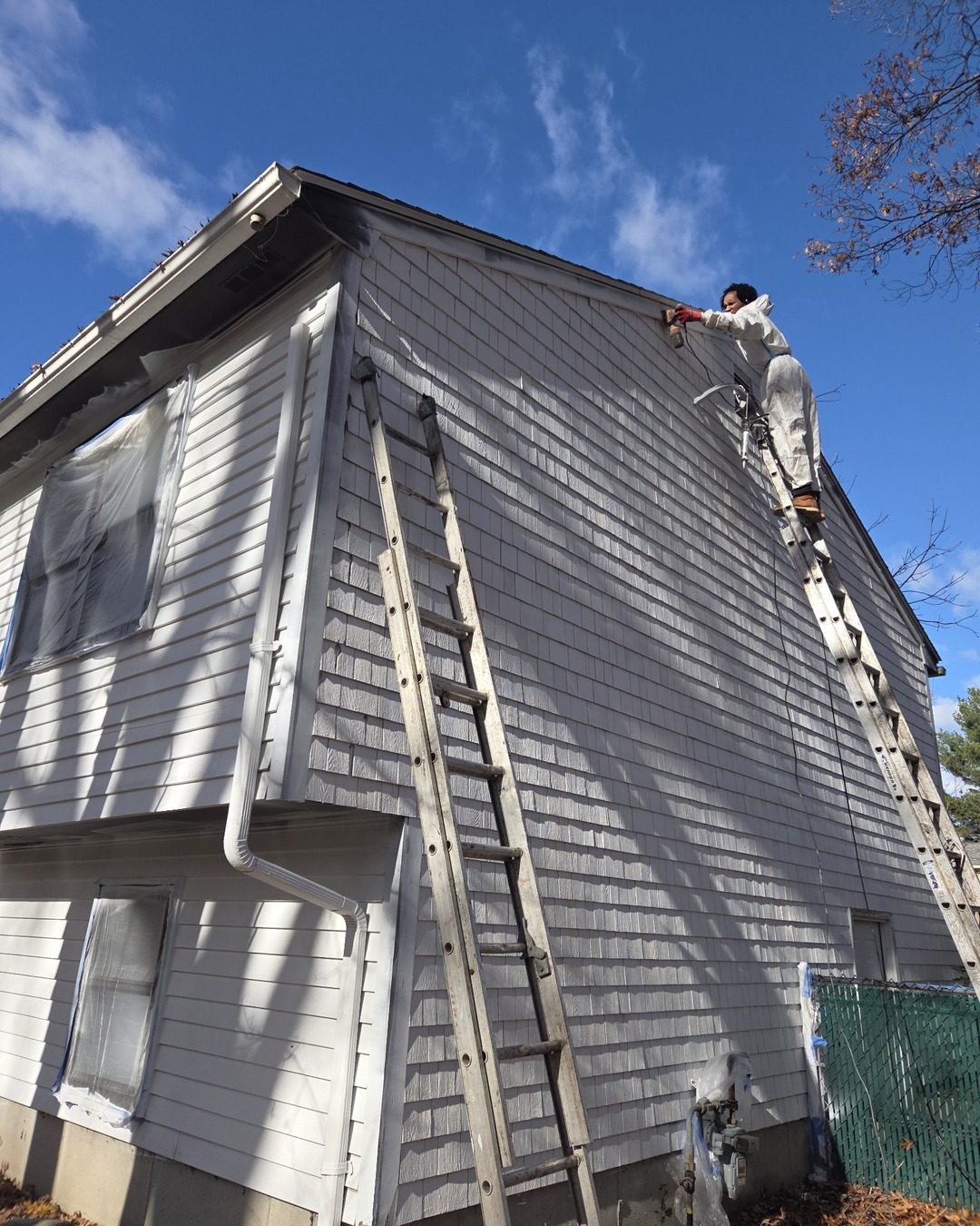 Two-story shingle home being spray-painted white, with tall extension ladders against the newly coated siding and windows masked off with plastic sheeting and tape