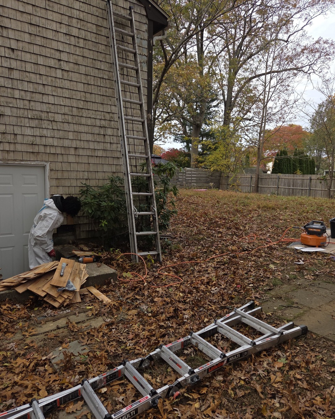 Basement entrance and lower corner of the shingle home during siding repair, with stacked replacement shingles, hand scrapers, an air hose and aluminum extension ladders