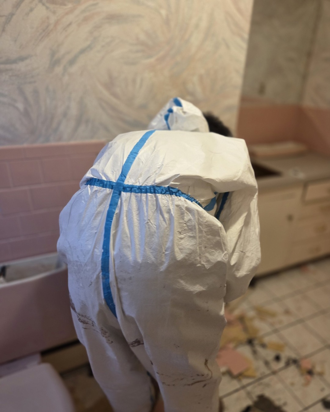 Worker in a protective suit clearing demolition debris from a bathroom with the original pink fixtures still in place